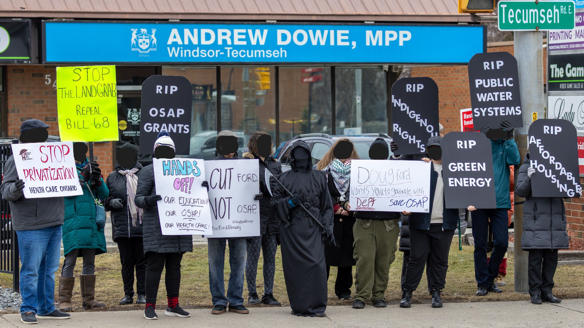 a line of people holding protest posters in front of MPP Andrew Dowie's office.